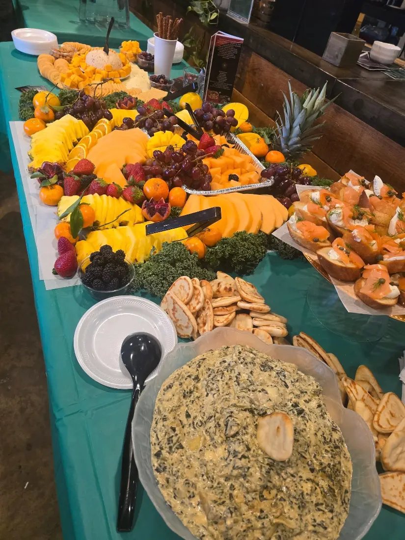Buffet line on a teal cloth with fruit trays, sandwiches and spinach dip in a bread bowl